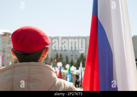 Military kid wearing red beret next to a Russian flag back view. Young patriotic boy in a uniform at a parade Stock Photo