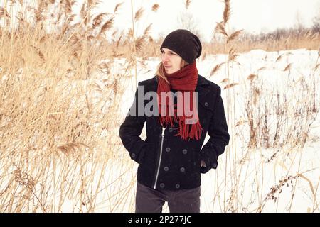 Young Caucasian man standing in a snow reed field alone, wearing a coat and a scarf. Sad blond male model winter portrait among dry grass.  White, bei Stock Photo