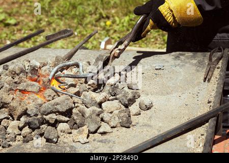 Blacksmith hand holding a metal swirl in tongs on the red hot glowing coals. Forge tools being used in an outdoor smithy Stock Photo