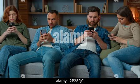 Focused four diverse african american caucasian hispanic friends sit on sofa in living room look at mobile screen together phone gadget internet Stock Photo