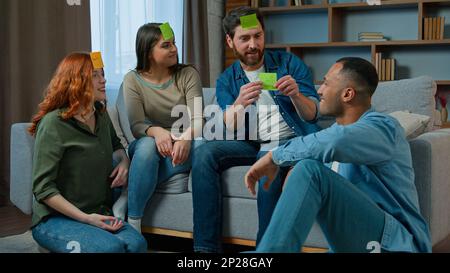 Diverse interracial friends women men playing guessing game with sticky notes on forehead play charades asking questions funny entertainment at home Stock Photo