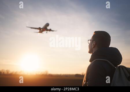 Man with backpack looking up to airplane landing at airport during beautiful sunset. Stock Photo