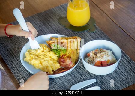 Kids menu, scrambled eggs, bacon, toast and muesli with yoghurt served on the restaurant table with plastic kids friendly eating utensil. Stock Photo