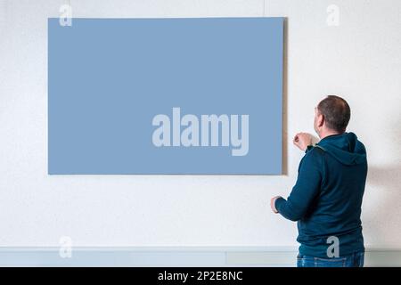 Upper body view of a man in casual clothing with his back to the camera adjusting a neutral image on a light colored wall. Stock Photo