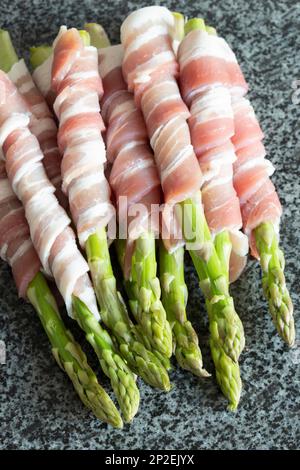 Raw organic asparagus, wrapped in streaky bacon, on a black granite worktop surface. Fresh seasonal produce concept Stock Photo