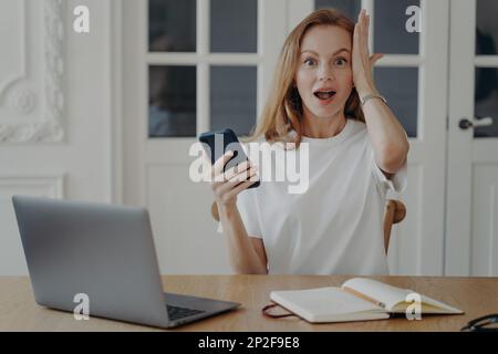 Emotional young woman holding smartphone looking at camera with opened mouth, surprised with online store offer. Amazed shocked female got unbelievabl Stock Photo