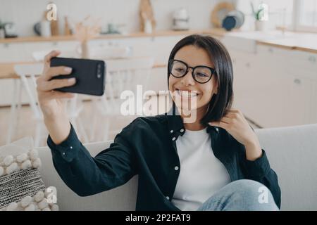 Smiling girl blogger holding smartphone, talks by video call, takes selfie photos, sitting on couch at home. Happy young woman wearing glasses chattin Stock Photo