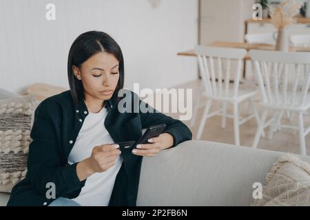 Focused female tenant holds bank credit card, phone, pay rental using online banking service at home. Serious woman makes money transfer in e-bank app Stock Photo