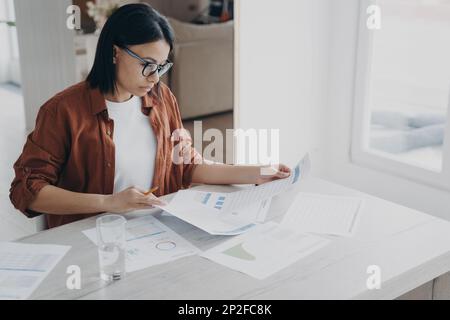 Focused businesswoman in glasses works on project, prepares report in charts analyzing documents at home office. Serious woman manager analyzes graphs Stock Photo