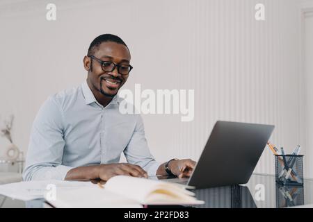 Pensive african american man wearing glasses working on laptop online, sitting at office desk, satisfied with his business project. Smiling black guy Stock Photo