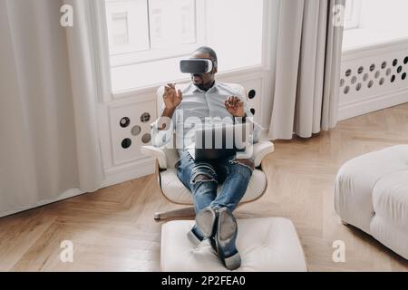 African american man in VR glasses working in augmented reality, sitting in armchair with laptop at home, pointing at virtual reality object, interact Stock Photo