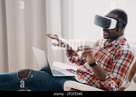 Smiling african american man in VR glasses sits in armchair with laptop, testing virtual reality goggles. Modern black guy viewing purchase in cybersp Stock Photo