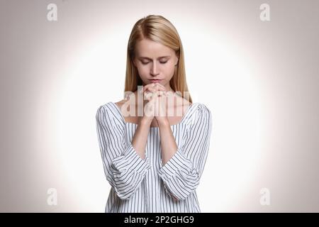 Religious young woman with clasped hands praying against light background Stock Photo