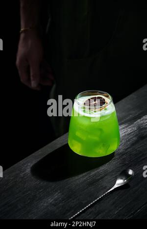 Special green cocktail of mixology, lemon rum, garnished with dried fruit, on black bar table, bartender in the background, clouse up Stock Photo