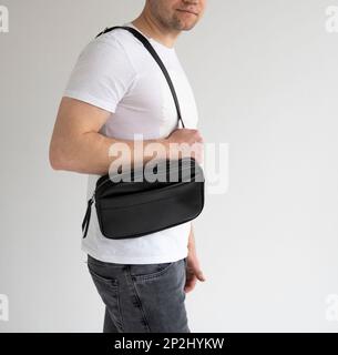 Side view man in a white t-shirt with a leather black handmade bag over his shoulder. Dark designer banana bag. Comfortable small bag for walking. Stock Photo