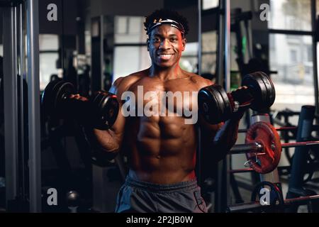 20s Black and muscular man in a gym showing dumbbells and his muscles with a smiling emotion Stock Photo