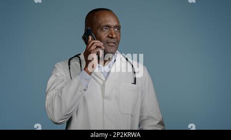 Mature african american man doctor talking on phone with patient in blue background studio male therapist in medical gown consult client using Stock Photo