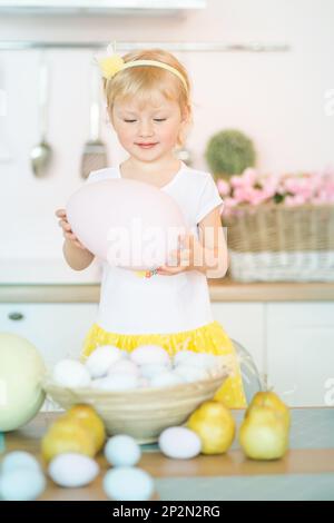 Cute little girl with a big pink Easter egg in the kitchen next to a basket of eggs. Easter children's portrait, funny emotions, surprise Stock Photo