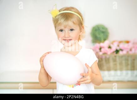 A smiling blonde girl is holding a large pink Easter egg in a bright kitchen with flowers. kid celebrates Easter Stock Photo