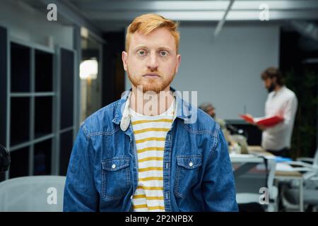 Young red-haired programmer or businessman in modern office looking seriously at camera. Stock Photo