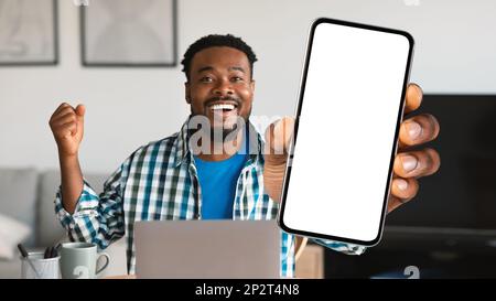 Excited Black Man Sitting At Desk With laptop And Showing Blank Smartphone Stock Photo