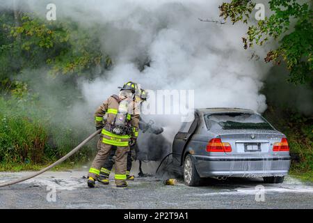 Saint John, NB, Canada - August 18, 2022: Smoke billows from a parked car while two firemen spray foam water into it. The firemen are wearing breathin Stock Photo
