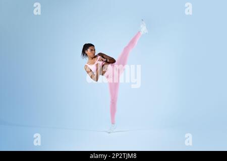 Determined black woman in sportswear doing high kick, tae-bo martial arts exercises on blue background, free space Stock Photo