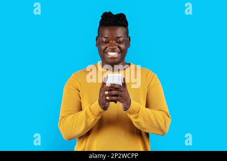 Young Black Man Using Modern Smartphone While Standing Over Blue Studio Background Stock Photo