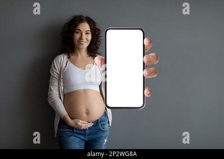 Happy smiling pregnant woman embracing belly and showing big blank cellphone Stock Photo