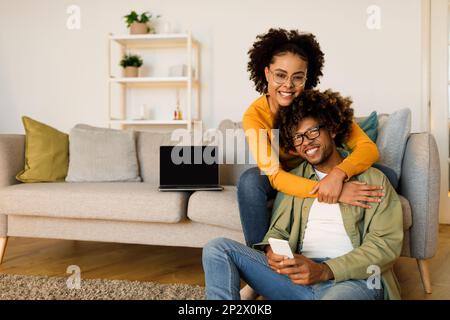 Happy Black Couple Hugging And Using Smartphone At Home Stock Photo