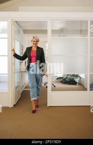 Successful businesswoman smiling at the camera while standing in front of her office. Happy female entrepreneur working in a creative workplace. Stock Photo