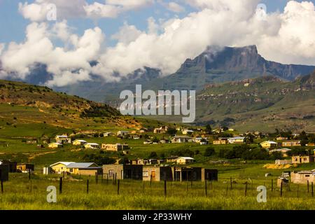 The majestic cliffs of the Drakensberg Mountains, of South Africa, shrouded in low-lying clouds with a small rural settlement in the foreground Stock Photo