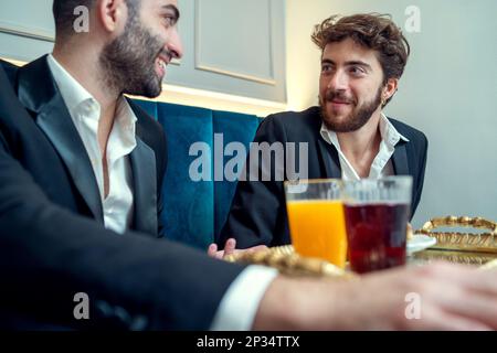 Two young men are sitting in a restaurant, enjoying breakfast and conversation. They are both dressed in jackets and shirts, and have glasses of fruit Stock Photo