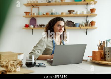 Potter entrepreneur using laptop in workshop, smiling Stock Photo