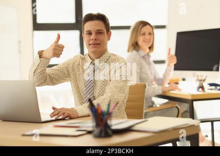 Two managers are sitting at their desks in office and showing thumbs up gesture. Stock Photo