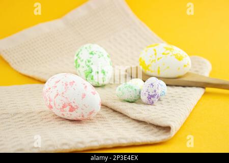 Composition of painted eggs, chicken and quail with a wooden spoon and a beige kitchen towel, on a yellow background.  Side view Stock Photo