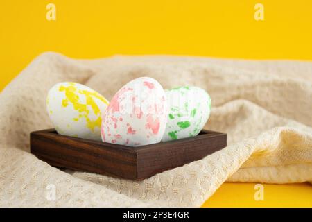 Easter-colored chicken eggs lie in a wooden low box, next to a kitchen towel lie on a yellow background Stock Photo