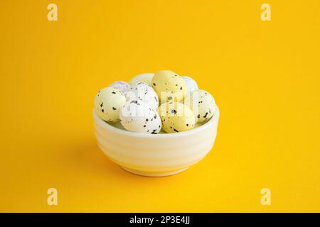 Several quail eggs lie in a white ceramic bowl on a yellow background.  Lots of empty space, Easter composition Stock Photo