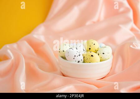 Several quail eggs lie in a white ceramic bowl on peach pink silk on a yellow background.  Lots of empty space, Easter composition Stock Photo