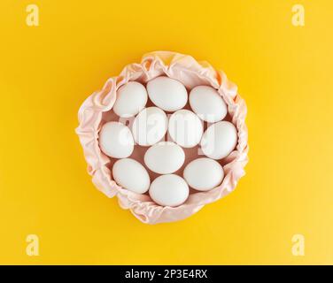 White chicken eggs lie in a pink silk basket on a yellow background.  Easter composition, lots of empty space Stock Photo