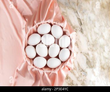 White chicken eggs lie in a basket covered with pink silk on a marble table.  Top view of easter composition, space for text Stock Photo