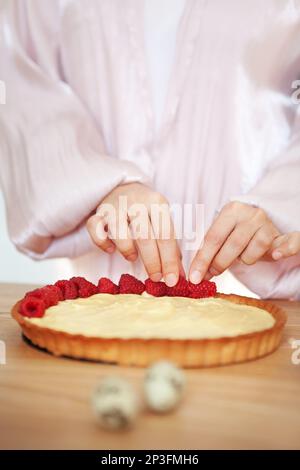 Chef Woman decorating cake with berries and cream cheese on kitchen table. Close-up of female hands making tartlet with fresh raspberries Stock Photo