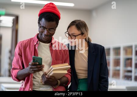 Two diverse students standing in classroom using smartphone during study break Stock Photo