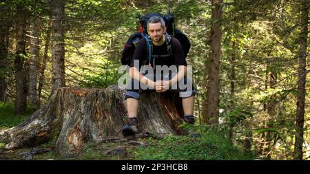 A man sitting with a backpack on a tree trunk by the trail. Dolomites, Italy Stock Photo