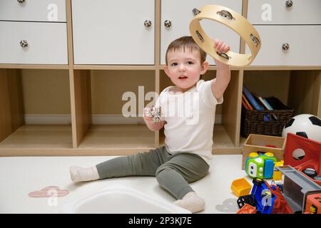 Toddler baby plays the tambourine while sitting on the floor in the children's room. Child boy playing musical instruments. Kid aged one year eight mo Stock Photo