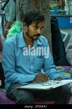 Kolkata, India - February 12, 2023: Middle aged male sketch artist sitting at a table, intently drawing a portrait with pencils on paper. Stock Photo