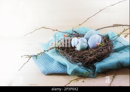 Blue and turquoise Easter eggs in a nest from twigs and quince branches with some flowers on cloth and a light wooden background, copy space, selected Stock Photo