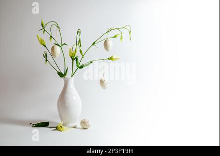 Delicate flower bunch in a white vase decorated with hanging Easter eggs on a light gray background, copy space, selected focus, narrow depth of field Stock Photo