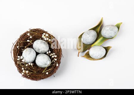 Brown nest of twigs with three gray Easter eggs, feathers, dried magnolia leaves and sprigs of gypsophila on gray background. Minimalistic Easter conc Stock Photo