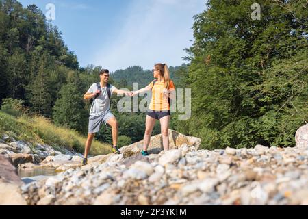 Young couple, walking along the rocky shore of a mountain river, exploring beautiful nature. Concept of an active vacation. Stock Photo